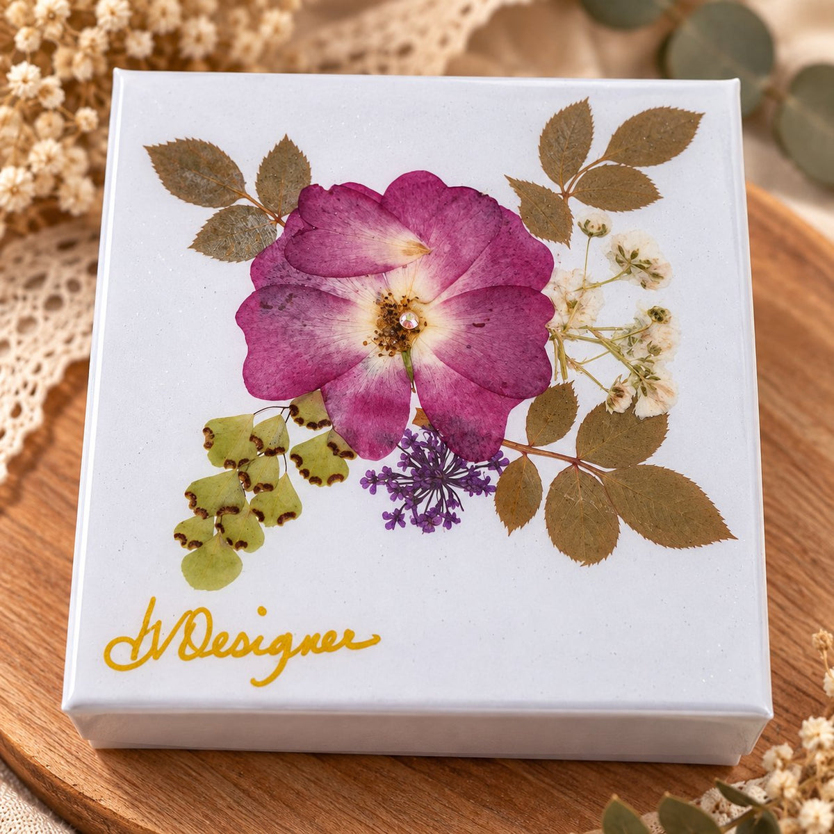 A square white gift box with a pressed floral design on the lid, featuring a prominent pink rose, green leaves, and small white blossoms. The box has a gold foil script logo reading 'ADesigner' and is photographed on a wooden surface with dried flowers in the background.