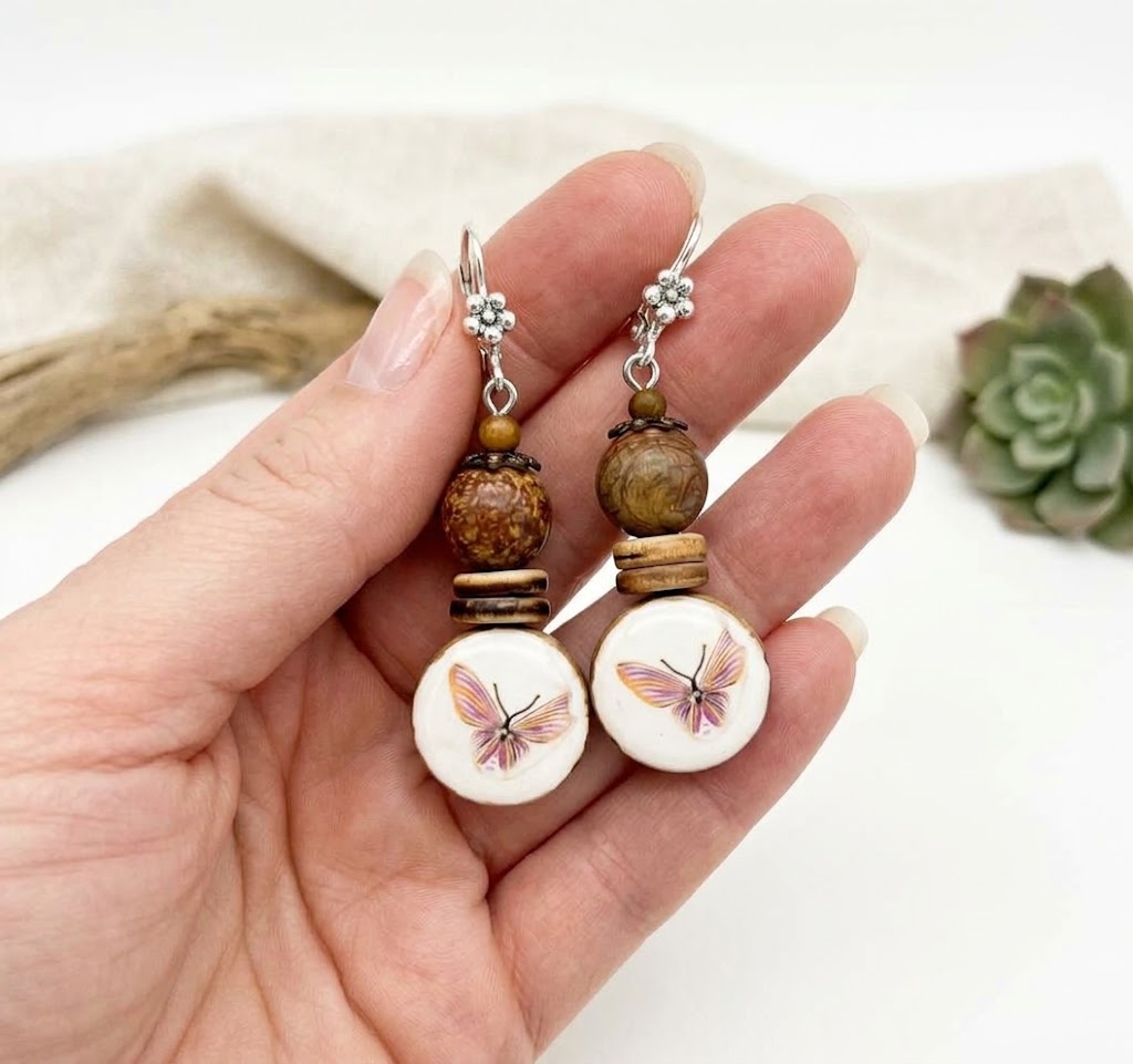 A pair of handmade dangle earrings held in a hand. The earrings feature silver-toned metal hooks with small floral charms, brown wooden beads, and round white disc charms printed with a pink and yellow butterfly.