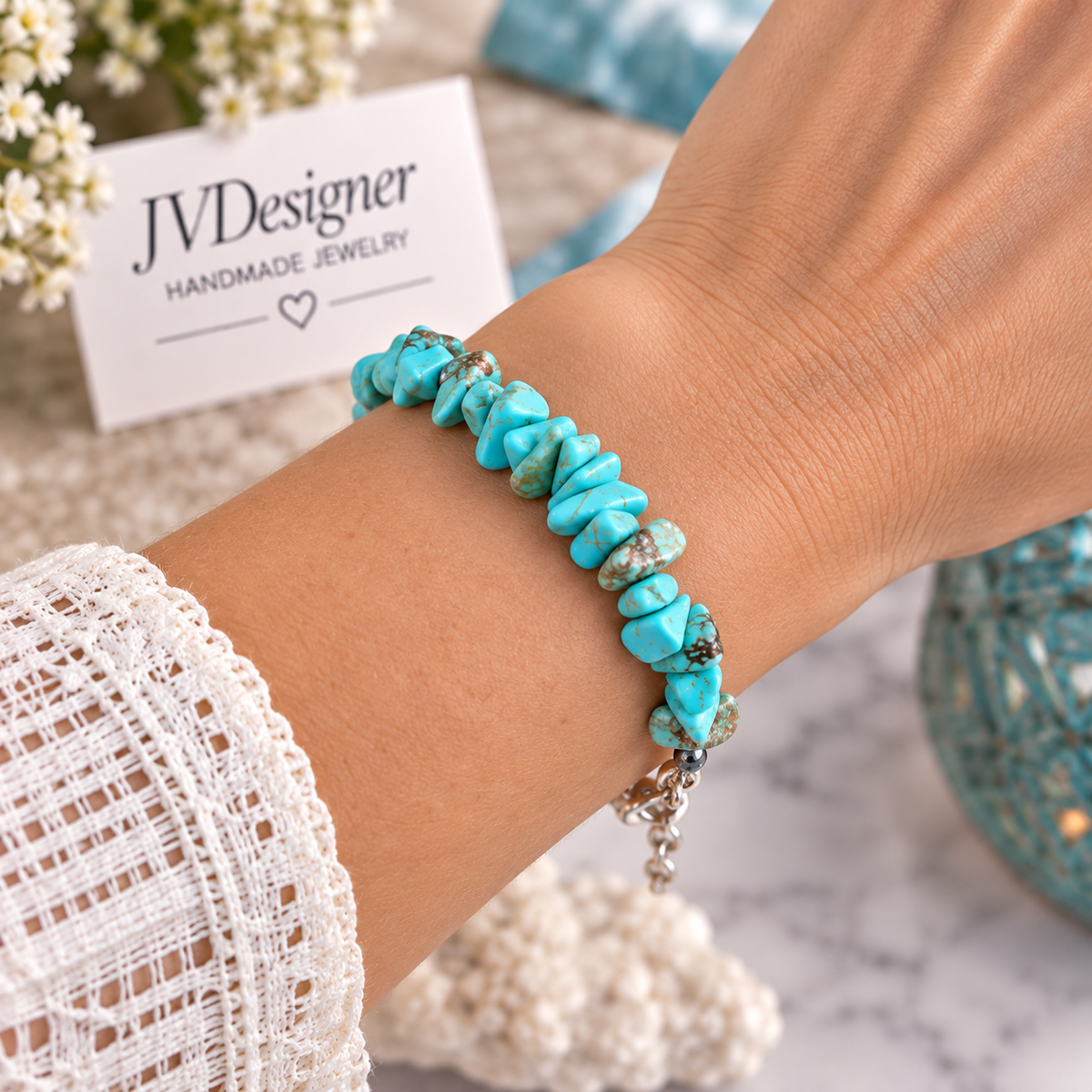 A close-up of a person's wrist wearing a handmade bracelet made of polished turquoise-colored stone chips with silver-toned metal links and a charm. A small card reading 'JVDesigner Handmade Jewelry' is visible in the background along with decorative flowers and a marble surface.