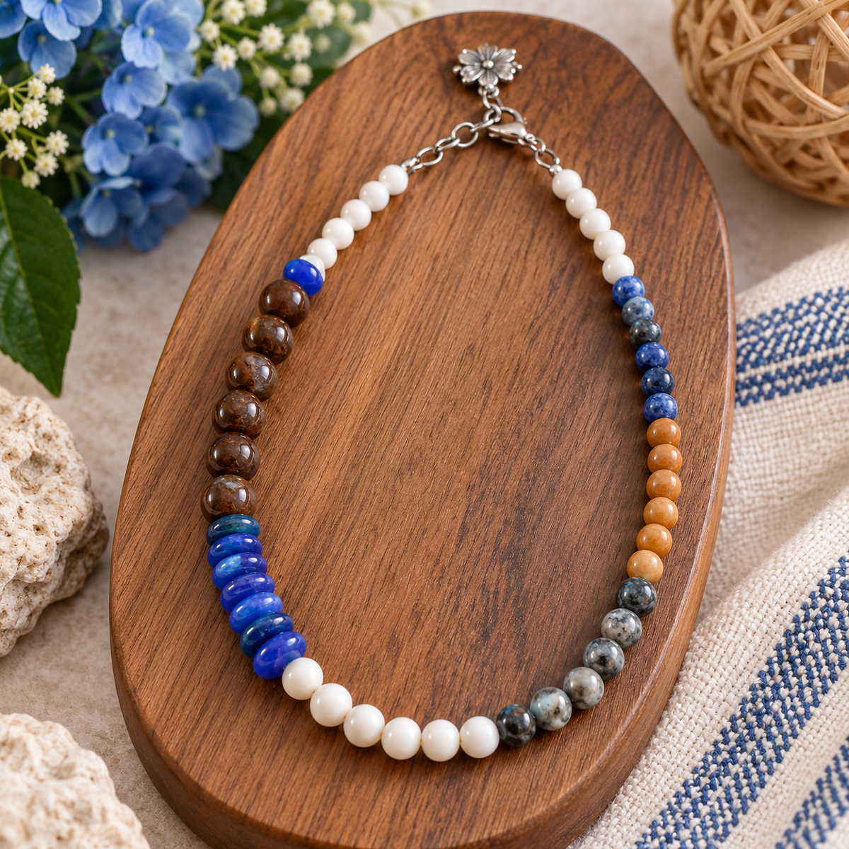 A beaded necklace with multiple distinct stone sections in blue, brown, white, orange, and gray, displayed on a carved wooden board with decorative flowers and fabric.