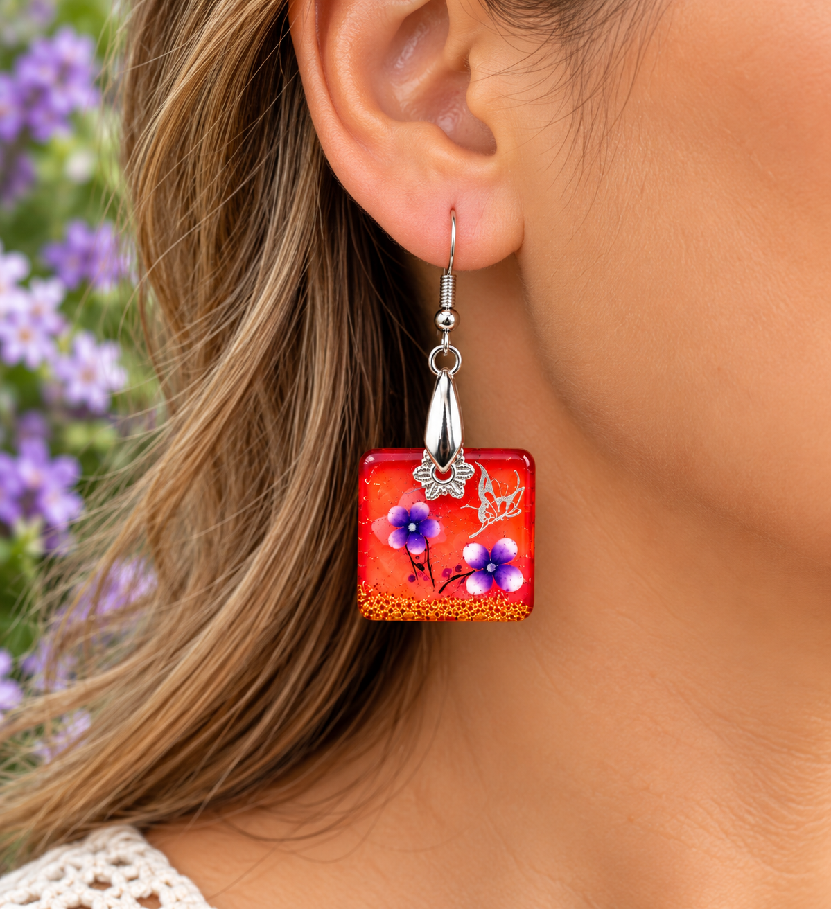 A close-up of a woman's ear wearing a square glass dangle earring. The earring is bright red-orange with hand-painted purple flowers, blue centers, and small orange details.