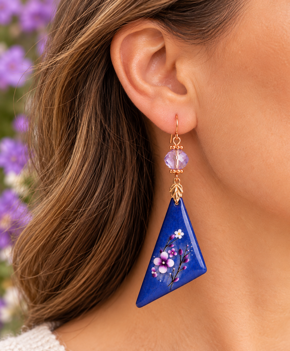 A close-up of a woman's ear wearing a dangling earring. The earring has a gold-tone hook, a round lavender bead, and a triangular blue pendant decorated with painted purple and white flowers.