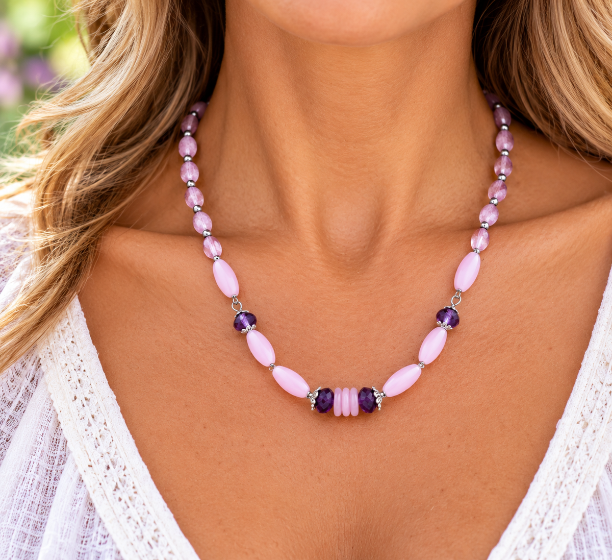 A close-up of a woman wearing a beaded necklace with light pink oval beads, purple faceted beads, and a central cluster of pink and purple beads, paired with a white lace top.