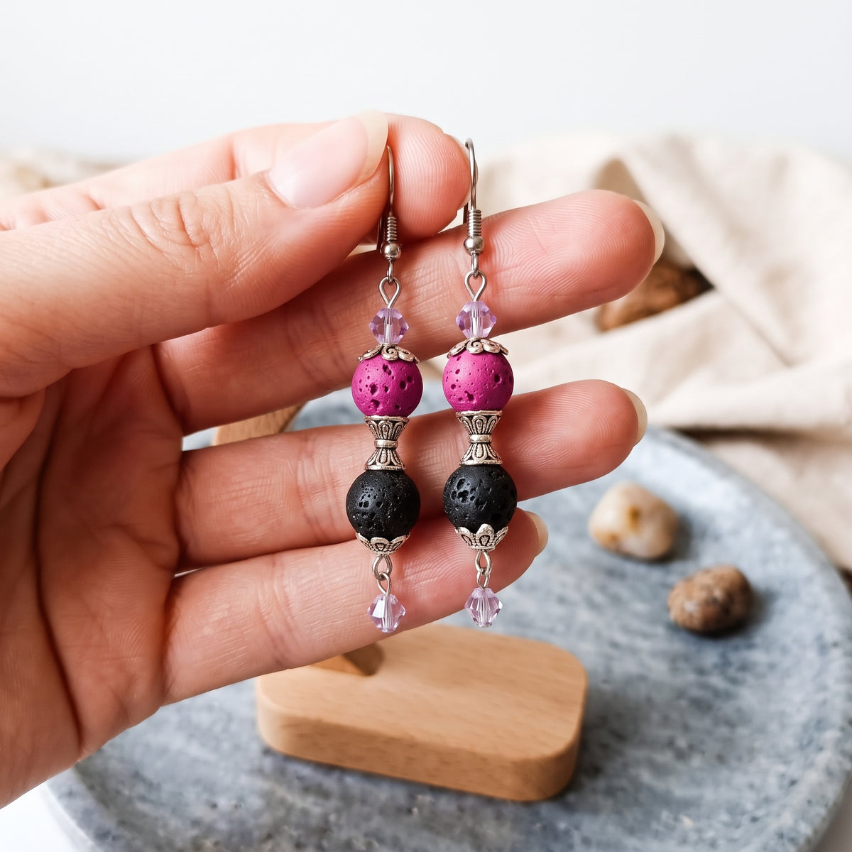 A close-up view of a hand holding the pink and black lava bead earrings with light purple crystals.