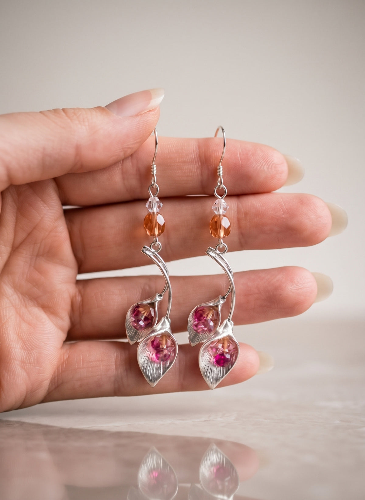 A hand holding a pair of silver dangle earrings with faceted orange beads and pink crystal leaf charms, set against a soft beige background.