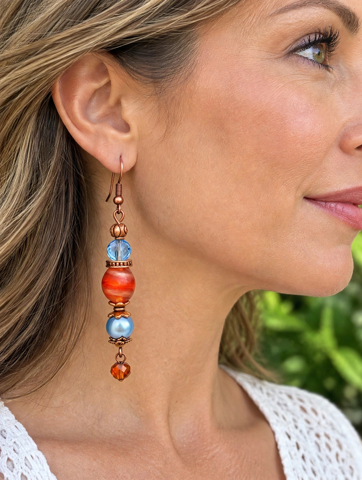 A close-up side profile of a woman wearing a long drop earring with copper-toned metal, light blue beads, and orange marbled beads, set against a blurred green outdoor background.