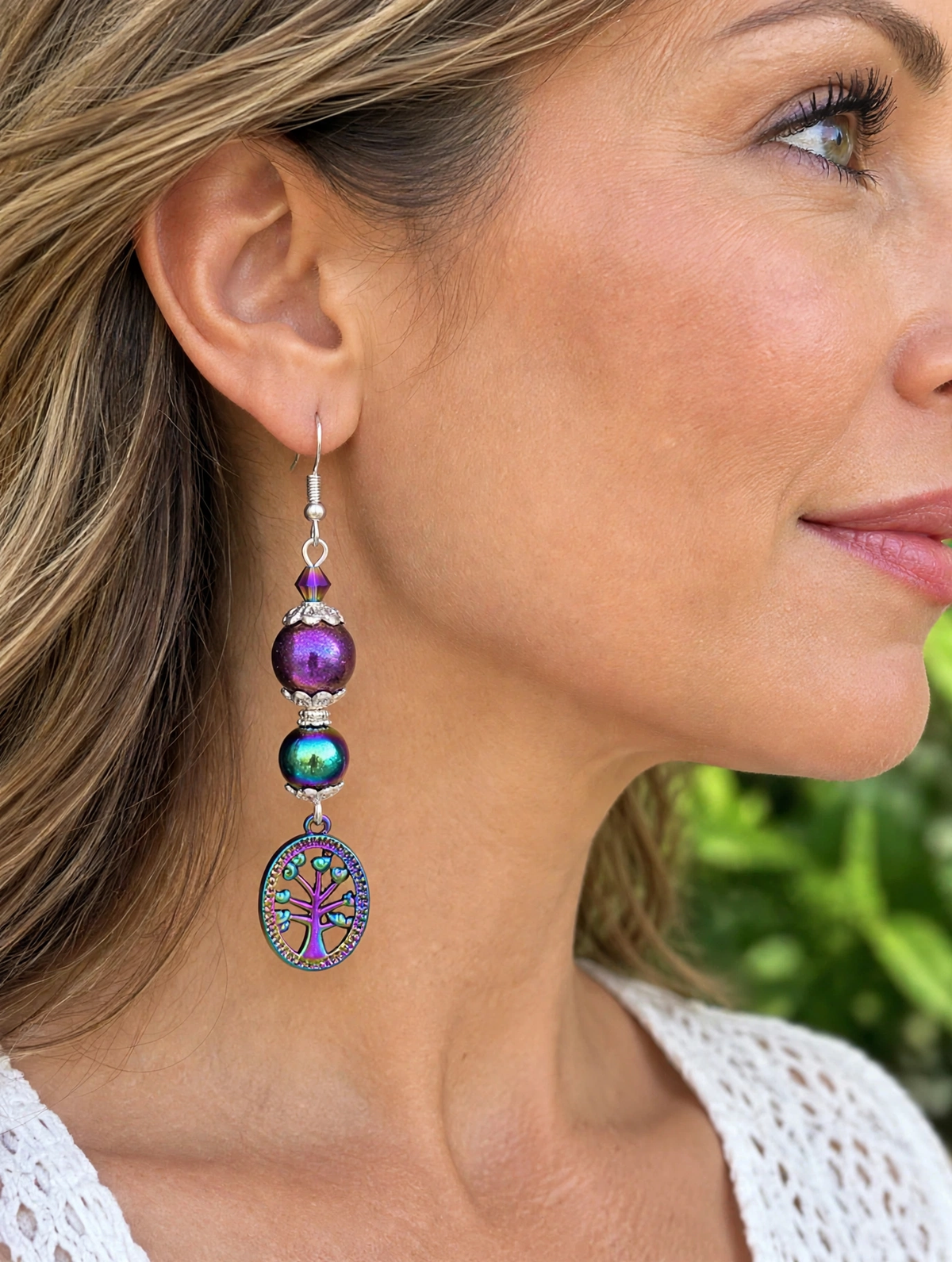A close-up side profile of a woman wearing a long dangling earring. The earring features a silver hook, a purple bead, an iridescent green bead, and a circular pendant with a colorful tree of life design.