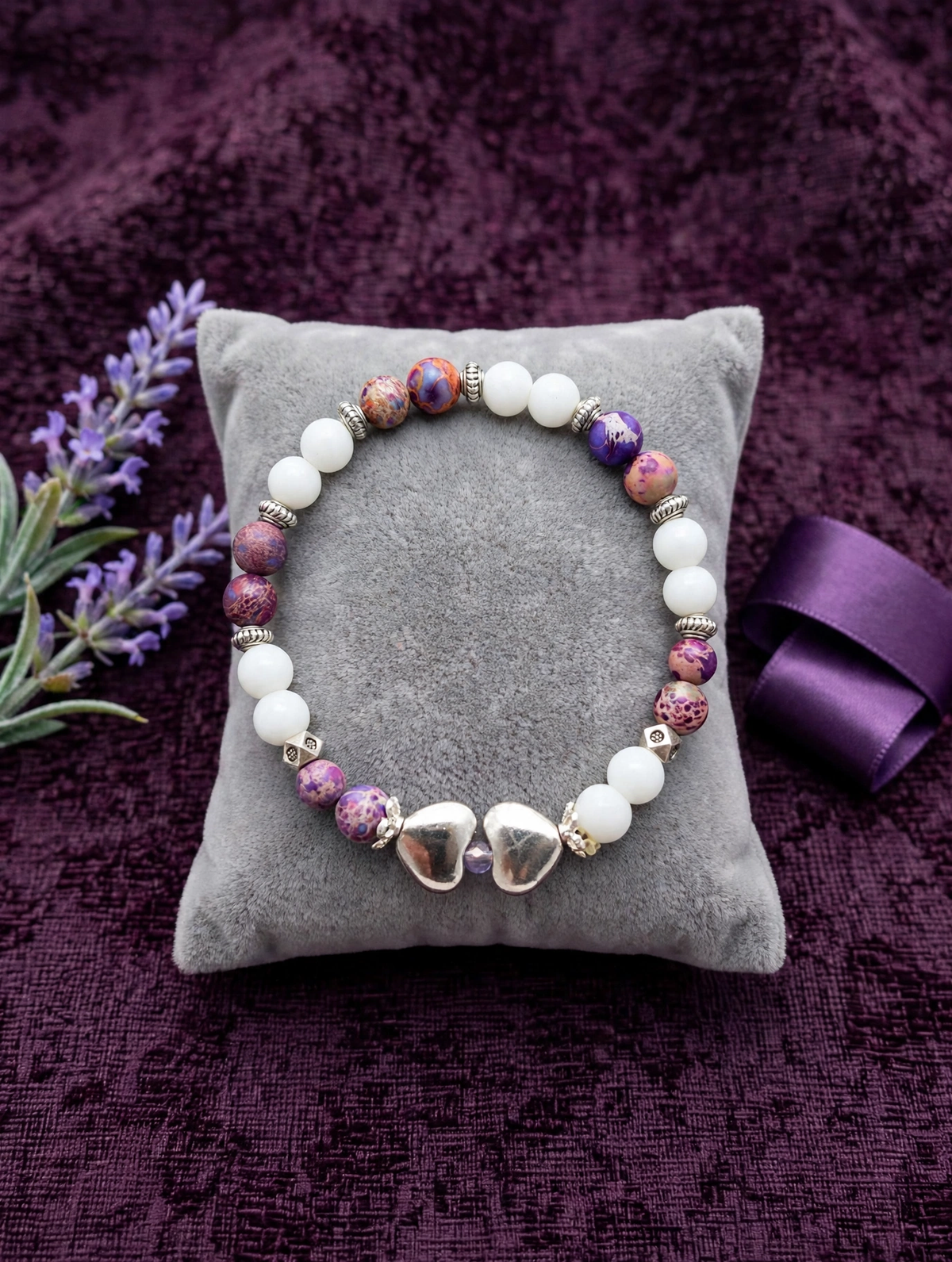 Stretch bracelet with white beads, purple speckled beads and silver spacer beads laid in a perfect circle, two silver hearts at the bottom with a light purple bead between them, on a grey velvet pillow over deep plum fabric with lavender flowers