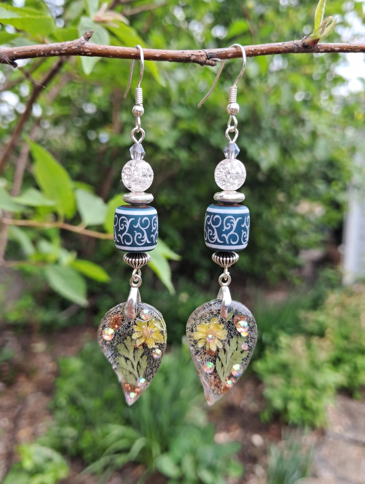 Handmade dried flower resin leaf dangle earrings with teal boho glass beads hanging from a tree branch
