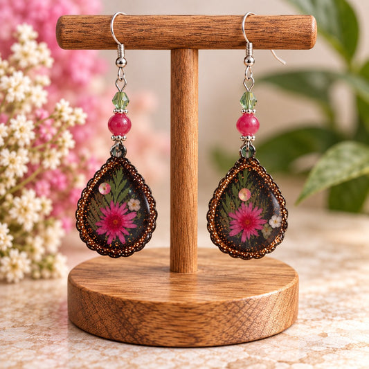Pink daisy resin teardrop earrings with bronze beaded edge, silver-tone fish hooks, and pink and light green bead accents, displayed on a wooden stand