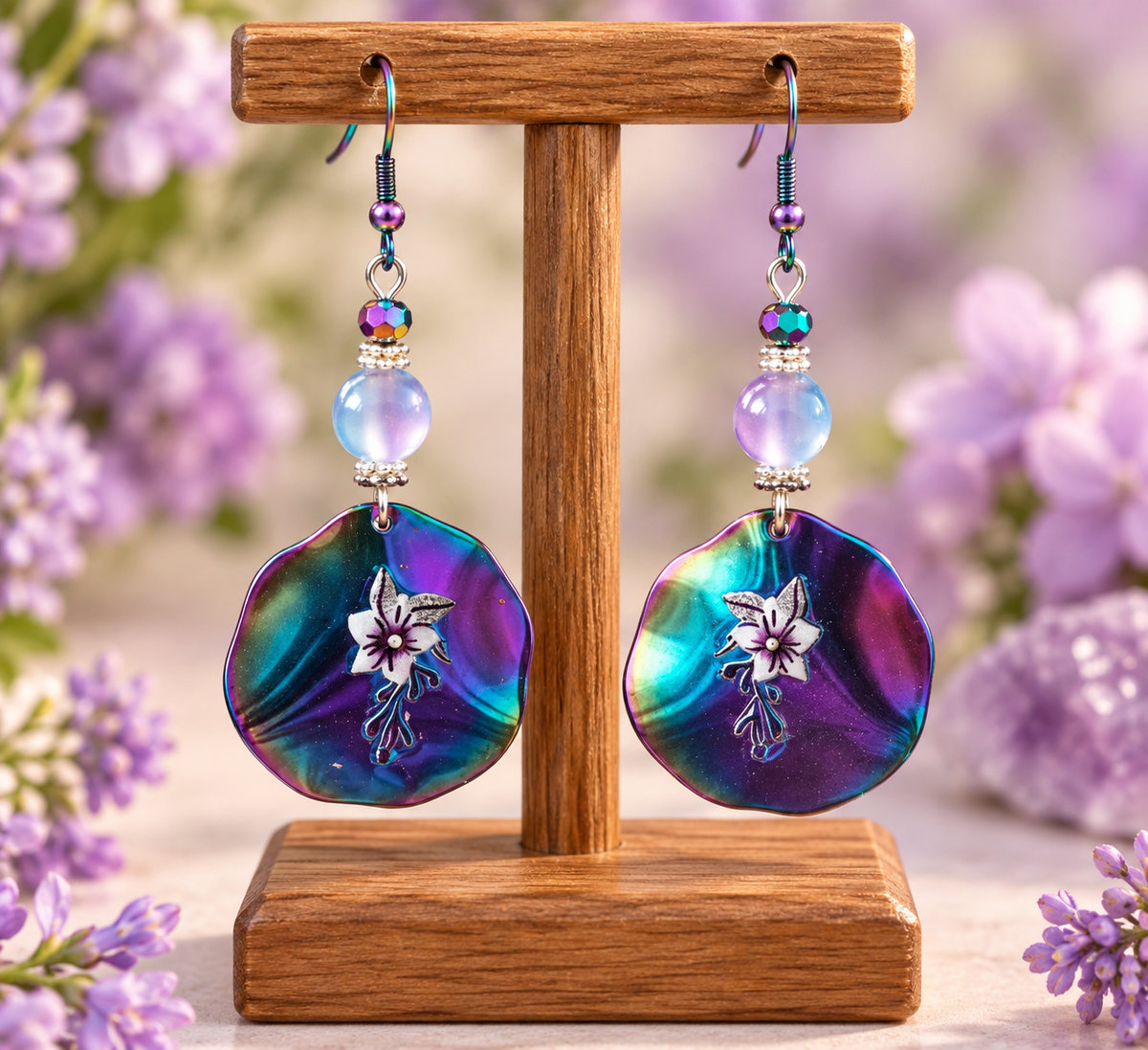 A pair of dangling earrings displayed on a wooden stand, featuring large iridescent shell-shaped dangles with a white flower motif and iridescent beads, set against a soft-focus background of purple flowers.