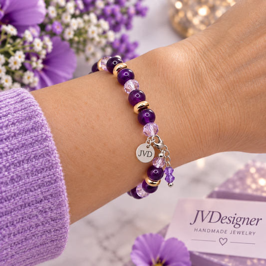 A close-up of a person's wrist wearing a beaded charm bracelet. The bracelet features purple beads, clear faceted beads, gold spacer beads, and a silver-tone charm engraved with 'JVD'.