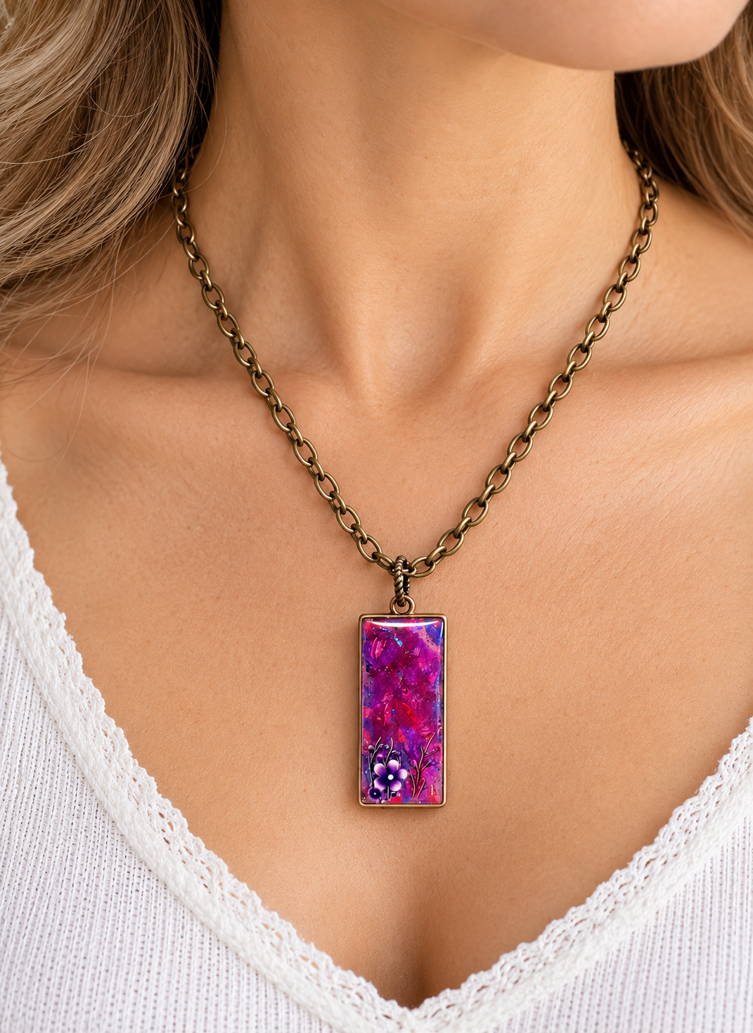 A close-up of a woman wearing an antique bronze chain necklace with a rectangular pendant featuring a vibrant purple and pink marbled pattern with small white floral details.