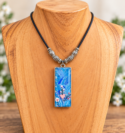 A rectangular pendant necklace with a vibrant blue abstract pattern, displayed on a wooden mannequin bust. The necklace features a black cord, textured silver spacer beads, and a silver-toned rectangular frame.
