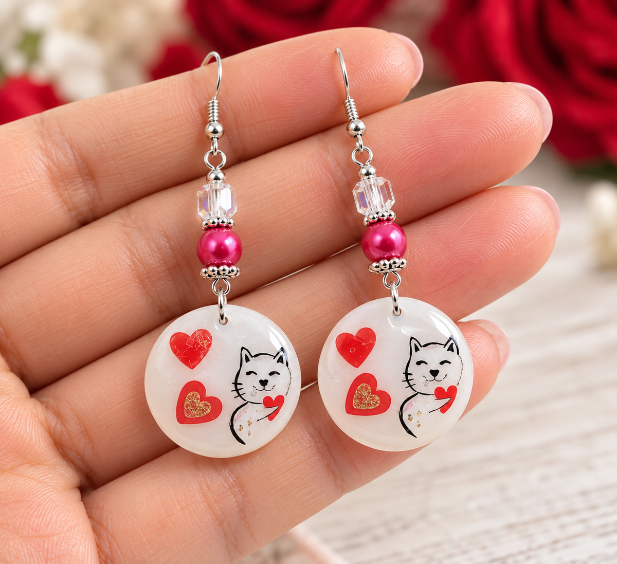 A pair of dangling earrings held in a hand. Each earring has a silver fishhook wire, a clear faceted bead, a bright pink bead, and a round white disc charm decorated with a black cat illustration, red hearts, and a small gold heart.