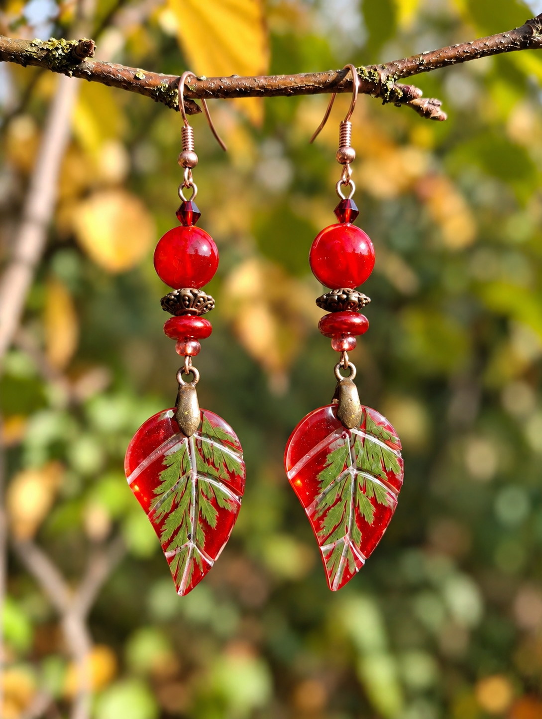 A pair of dangling earrings hanging from a tree branch outdoors. Each earring has copper-toned hooks, red beads, small metallic spacer beads, and a red glass leaf charm with green leaf vein details, against a blurred autumn foliage background.