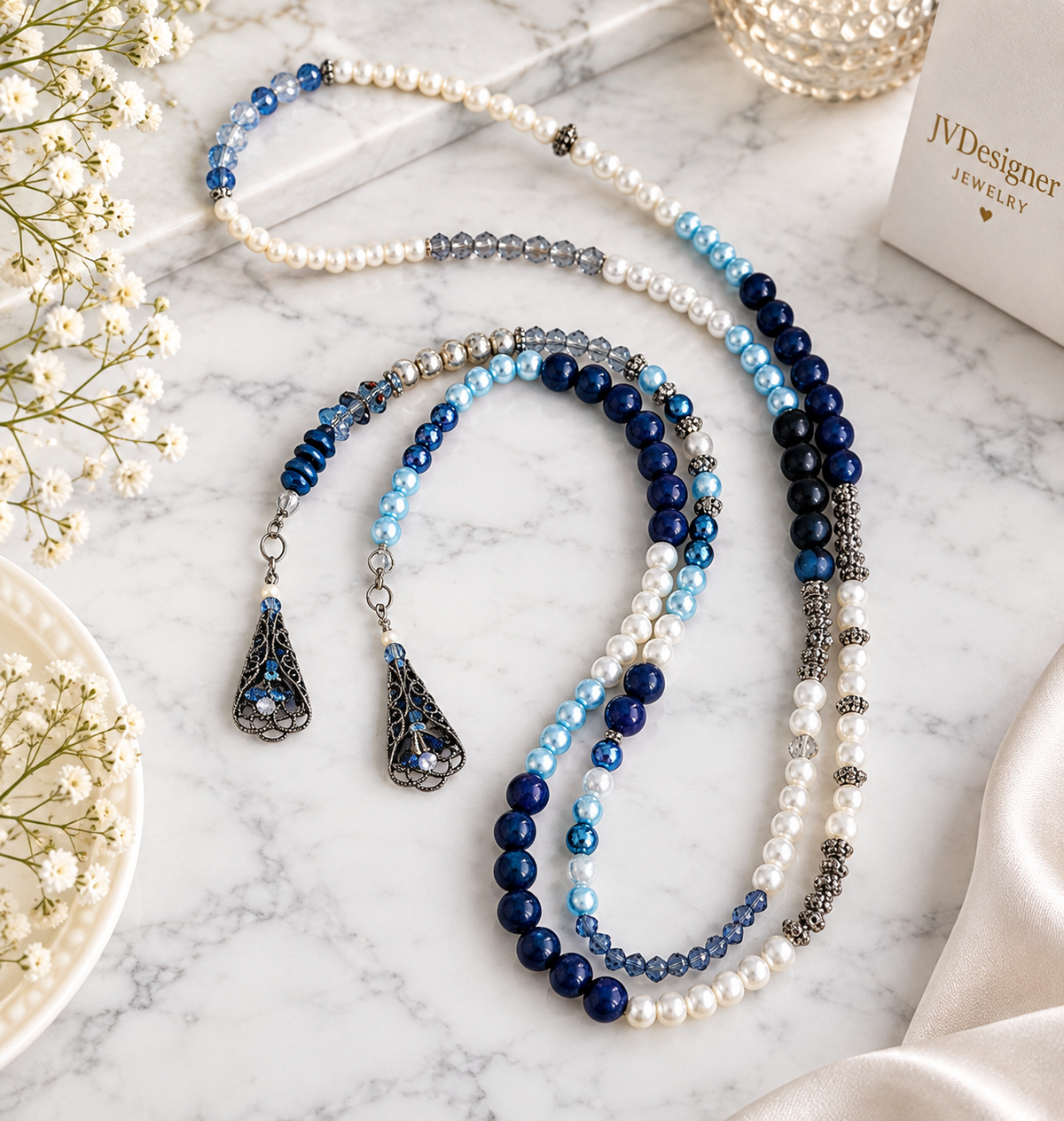A beaded necklace set with white pearls, light blue beads, dark blue beads, and silver spacer beads, styled on a white marble surface with baby's breath flowers and a jewelry box in the background.