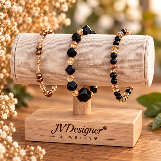 Three beaded bracelets displayed on a cylindrical fabric jewelry stand mounted on a wooden base branded JVDesigner Jewelry. The bracelets feature black faceted beads and metallic gold and clear beads, set against a softly blurred warm-toned floral background.