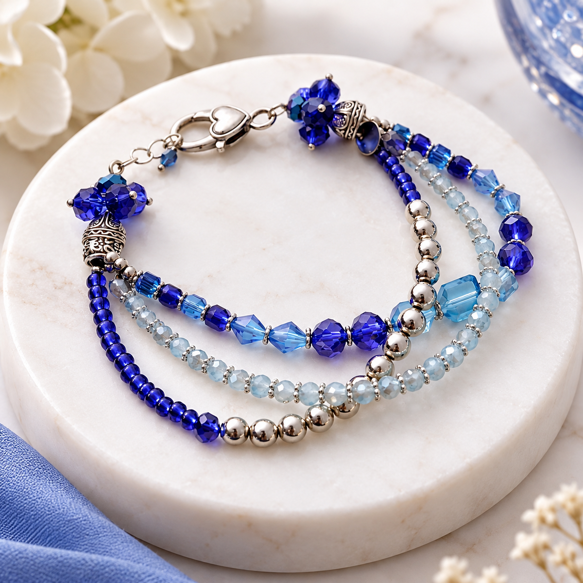 A beaded bracelet displayed on a round white marble stand. The bracelet features three strands: one with deep royal blue beads, one with light blue faceted beads and silver-toned metal beads, and one with silver-toned beads.