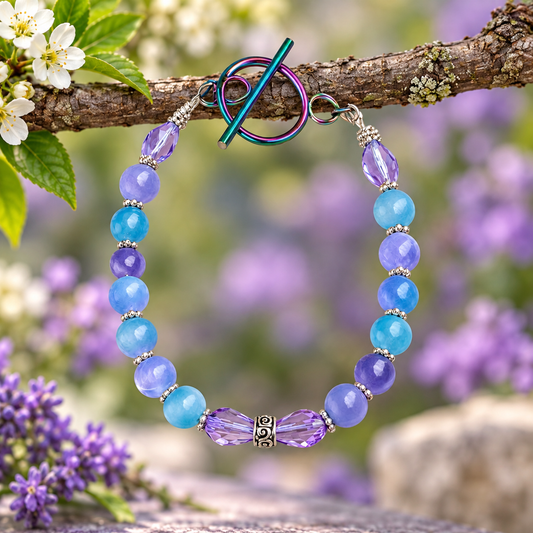 A beaded bracelet with light blue and lavender purple faceted beads, silver spacer beads, and a metallic toggle clasp, hanging from a tree branch with blurred purple flowers in the background.