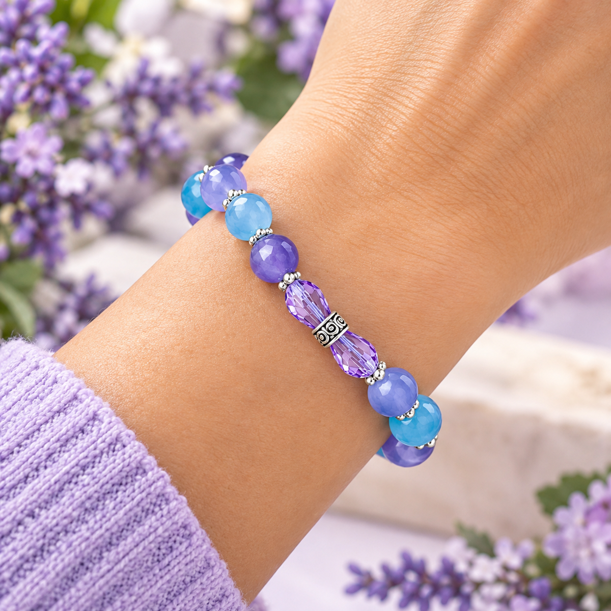 A close-up of a person's wrist wearing a beaded bracelet with light blue and purple beads, a faceted purple centerpiece, and a silver-toned decorative accent, set against a soft background of purple flowers.