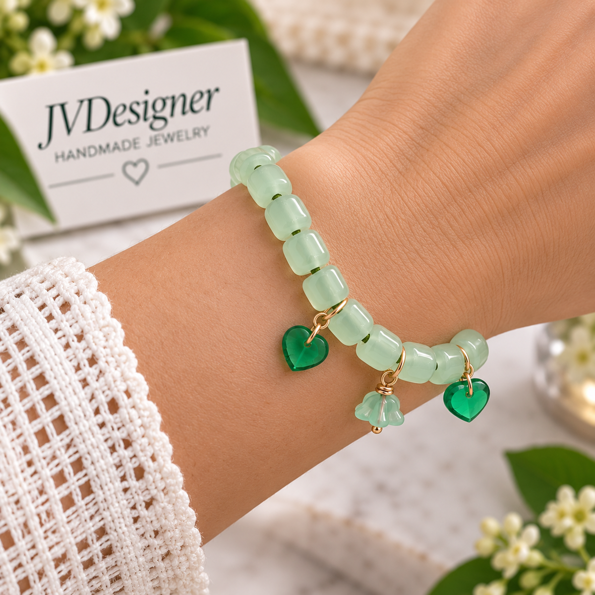 A close-up of a person's wrist wearing a handmade beaded bracelet. The bracelet features light green translucent rectangular beads, two green heart-shaped charms, and one small green flower charm with gold-tone metal links.