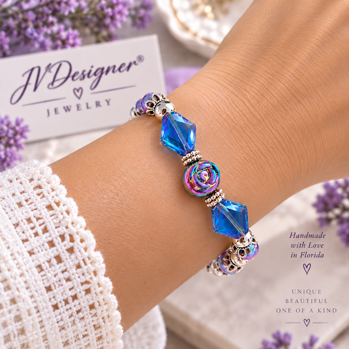 A close-up of a person's wrist wearing a handmade beaded bracelet with blue faceted beads, a rose-shaped bead, and silver-toned spacer details. The background includes a jewelry box labeled 'JVDesigner Jewelry' and decorative purple flowers.