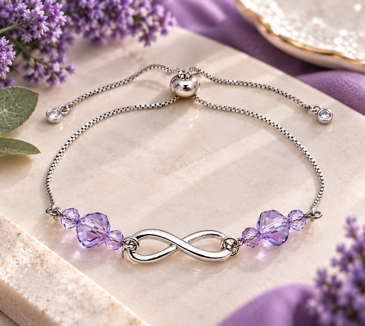 A silver infinity bracelet with faceted purple crystal beads, displayed on a beige stone slab with purple flowers and a purple fabric background.