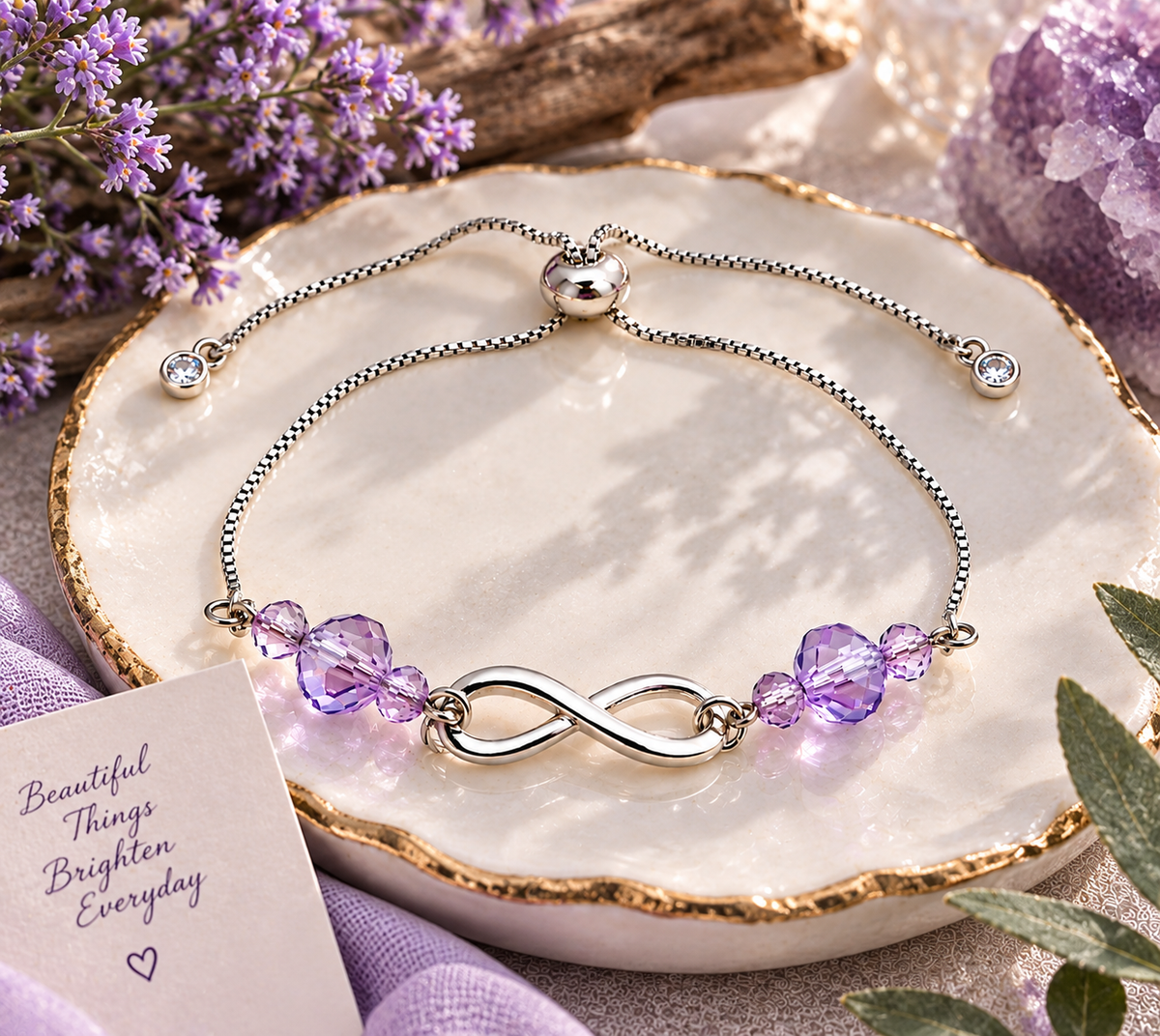 Silver infinity bracelet with sparkling lavender crystal beads and adjustable silver slider chain, displayed on an elegant cream and gold ceramic dish with purple flowers.