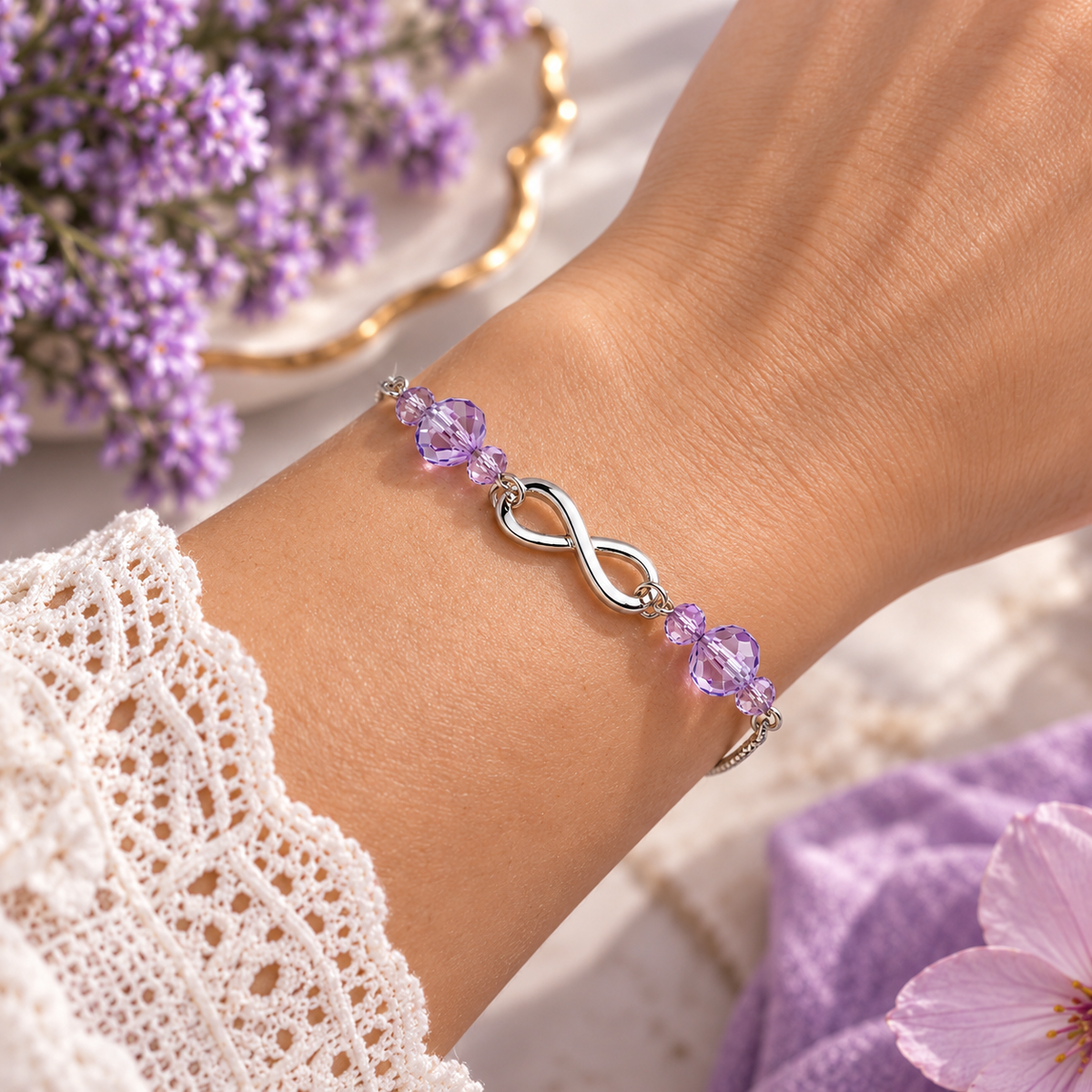 A silver infinity bracelet with purple crystal beads, shown worn on a woman's wrist. The background features soft purple flowers, a white lace sleeve, and a purple knit fabric.