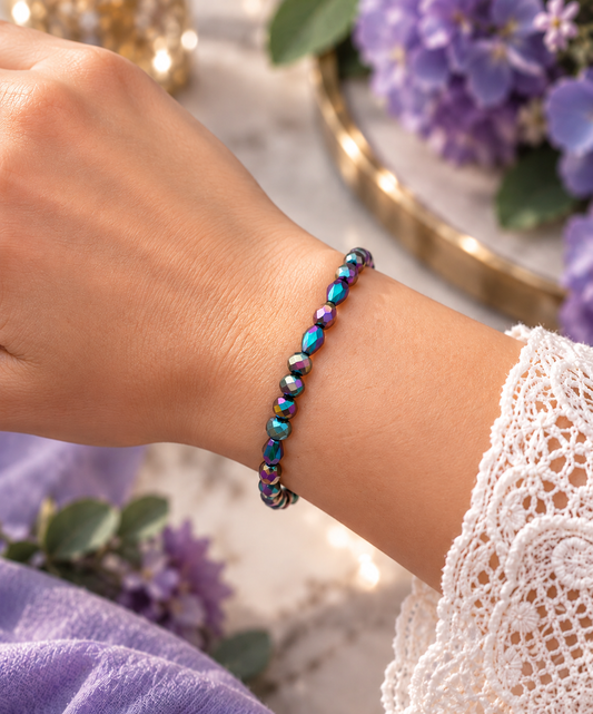 A close-up of a person's wrist wearing a beaded bracelet with iridescent blue, purple, and green beads. The person is wearing a white lace sleeve, with purple flowers and a gold tray blurred in the background.