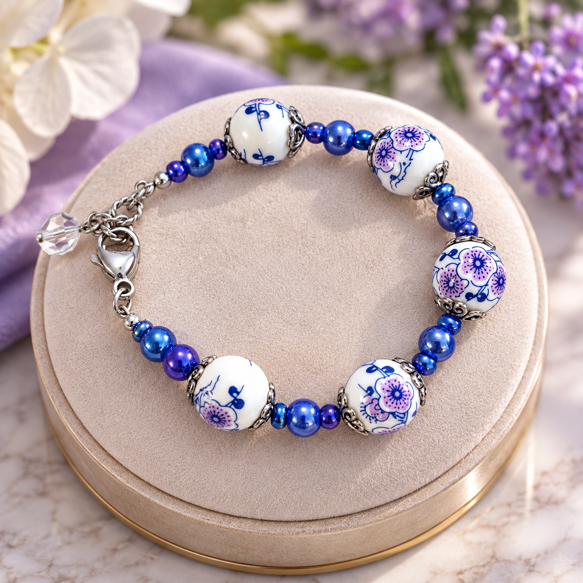 Blue and white floral ceramic bead bracelet with cobalt blue and purple beads, silver accents, and a crystal charm displayed on a cream velvet jewelry cushion.