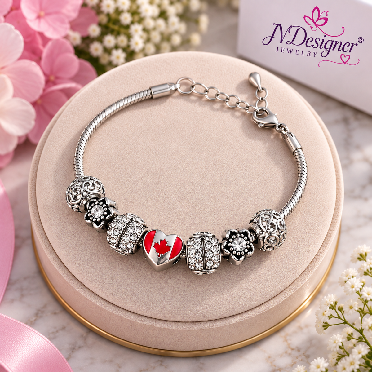 A silver-toned charm bracelet displayed on a round beige jewelry stand. The bracelet features a heart-shaped charm with a red maple leaf, flanked by crystal-encrusted and floral charms.