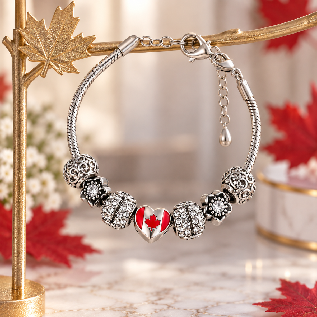 A silver-toned snake-style charm bracelet displayed on a gold branch stand. The bracelet features five decorative beads, including a central heart charm with a red maple leaf on a white background, and clear rhinestone accents.