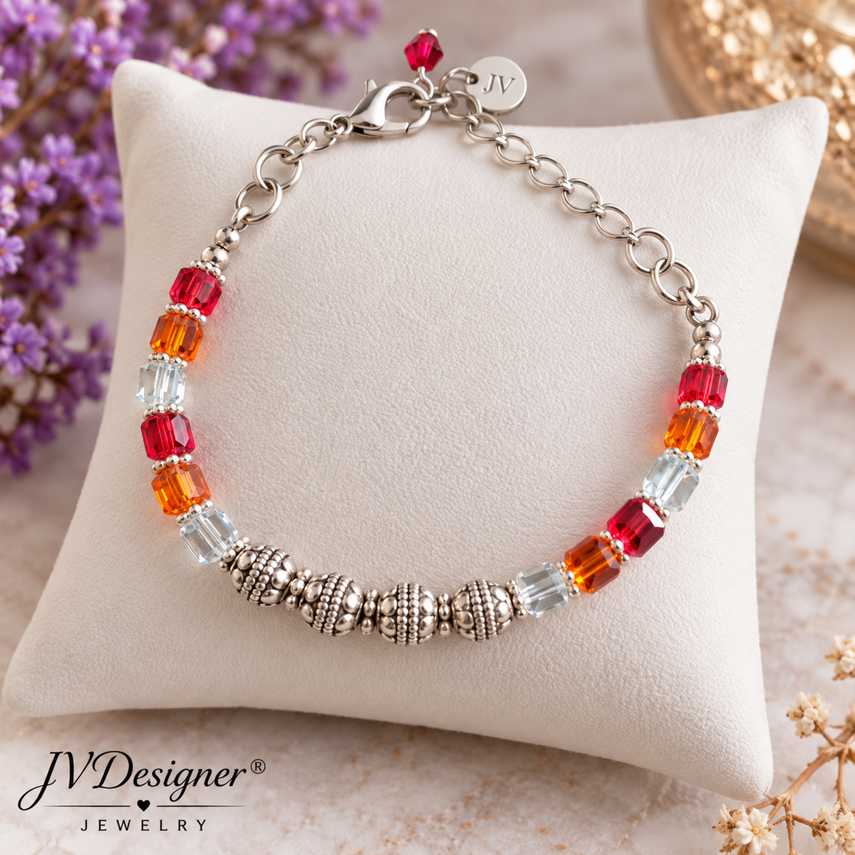 A silver chain bracelet with faceted beads in red, orange, and clear crystal, displayed on a cream-colored cushion. The brand logo 'JVDesigner Jewelry' is visible in the bottom-left corner.