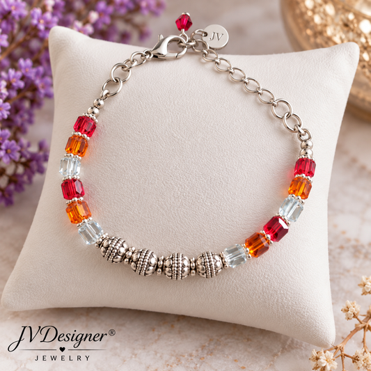 A silver chain bracelet with faceted beads in red, orange, and clear crystal, displayed on a cream-colored cushion. The brand logo 'JVDesigner Jewelry' is visible in the bottom-left corner.