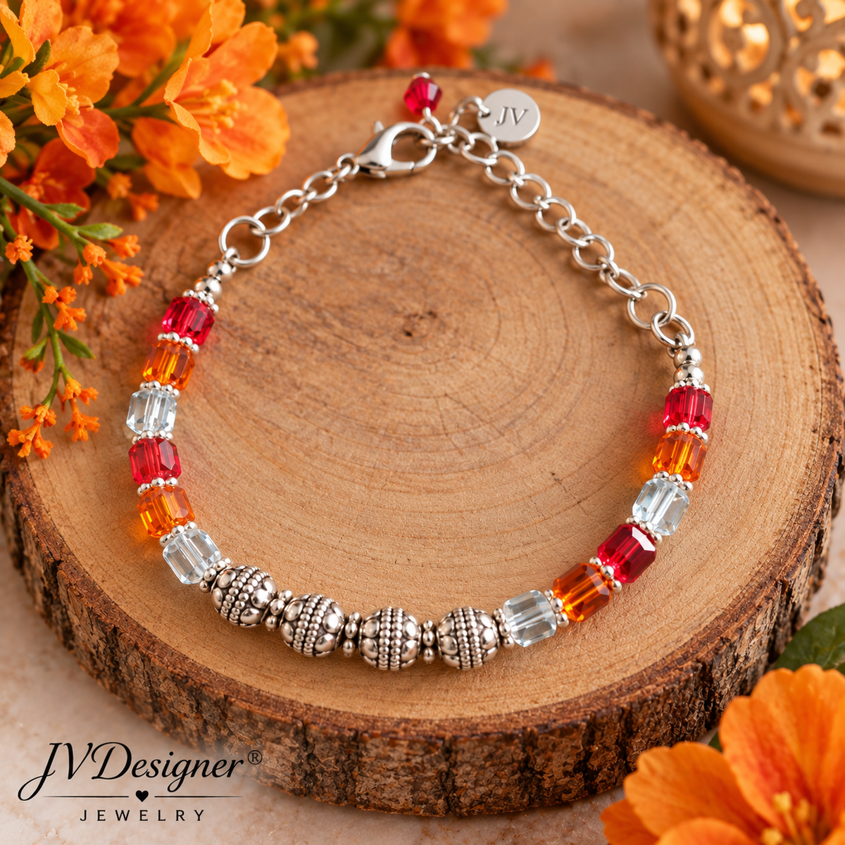 A silver chain bracelet with faceted red, orange, and clear beads, displayed on a rustic wood slice with orange flowers. The brand logo 'JVDesigner Jewelry' is visible in the bottom left corner.