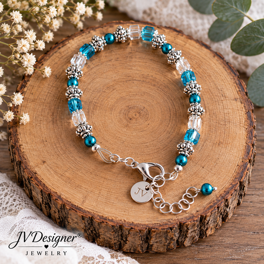 A beaded bracelet with turquoise blue beads, clear faceted beads, and silver-toned spacer beads, displayed on a round wood slice. The bracelet has a lobster clasp, extender chain, and a round initial charm engraved with 'M'.
