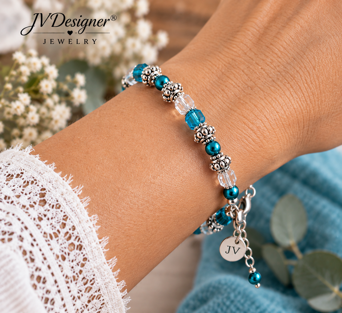 A close-up of a person's wrist wearing a beaded bracelet with turquoise blue beads, silver-toned spacer beads, clear faceted beads, and a dangling silver charm engraved with 'JV'. The background includes soft white flowers, green leaves, and a blue textured fabric, with the brand name 'JV Designer Jewelry' visible in the top left.