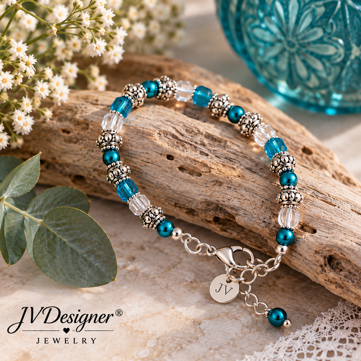 A beaded bracelet with turquoise blue beads, silver filigree spacers, and clear faceted beads, displayed on a piece of driftwood with eucalyptus leaves and white flowers. The bracelet has a lobster clasp and a charm engraved with 'JV'.