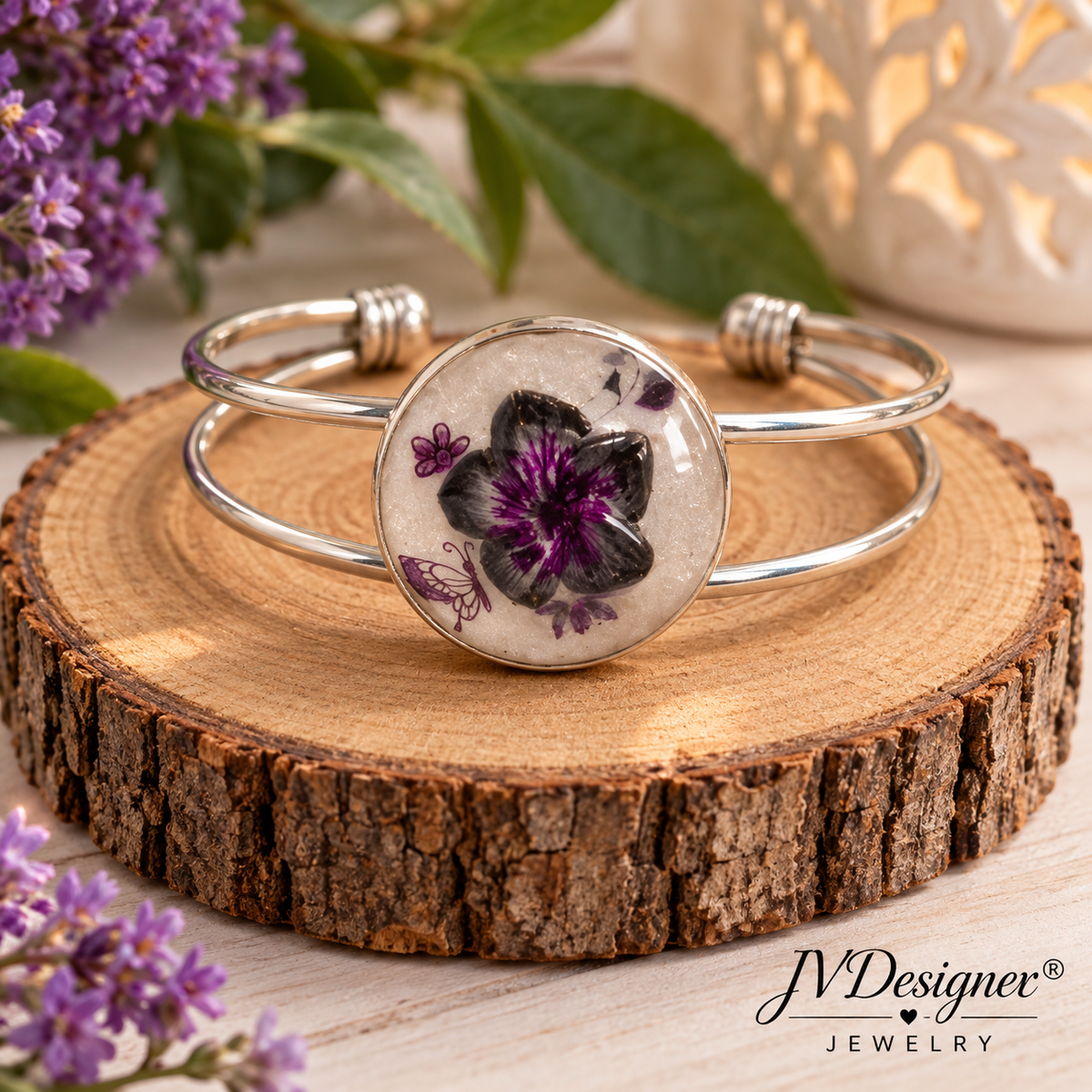 A silver-toned open cuff bracelet with a round clear resin cabochon featuring a black and purple flower design, displayed on a rustic wood slice with purple flowers and green leaves in the background. The brand name 'J'Designer Jewelry' is visible in the bottom right corner.