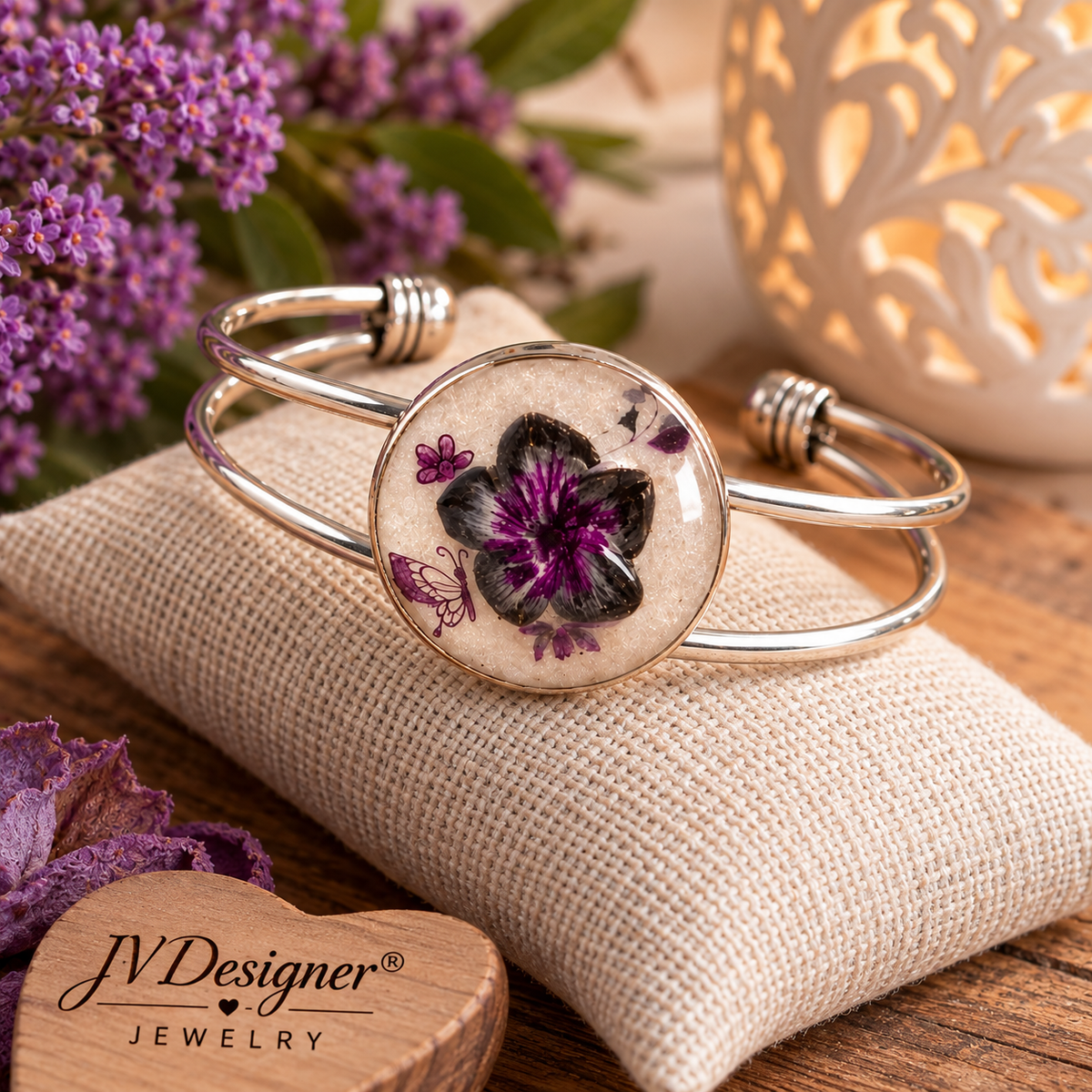 A silver-toned open cuff bracelet with a round clear-domed centerpiece featuring a black and purple flower on a beige background, styled on a burlap cushion with purple flowers and a wooden brand plaque reading JVDesigner Jewelry.