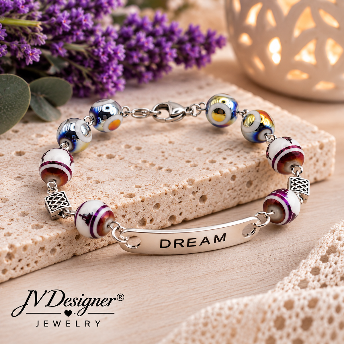 A silver charm bracelet with colorful glass beads and a central bar engraved with 'DREAM', displayed on a textured stone block with purple flowers and a warm glowing ceramic lantern in the background. The brand name 'JVDesigner' is visible in the bottom left corner.