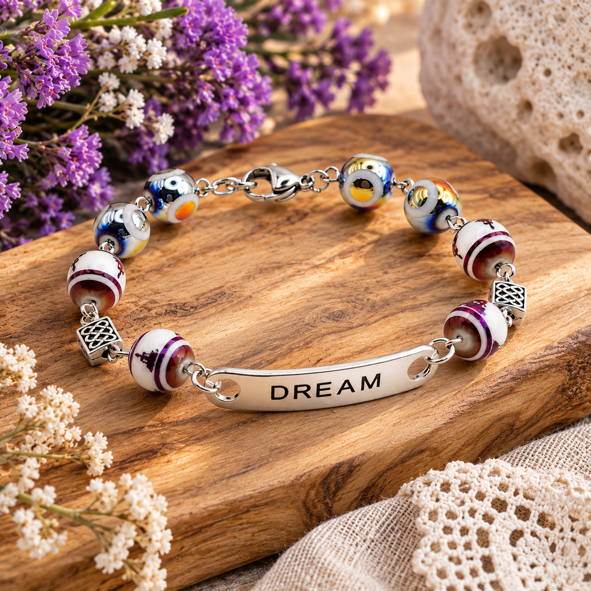 A silver-toned beaded bracelet with a central engraved bar reading “DREAM,” displayed on a wooden surface with dried flowers and textured decor in the background.