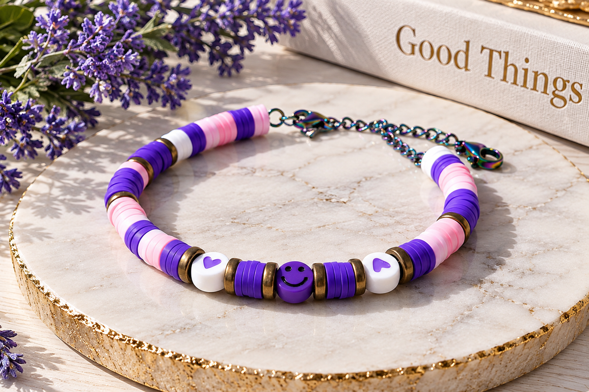A beaded bracelet with purple, pink, and white beads, a purple smiley face charm, and white heart charms, displayed on a round marble slab with decorative flowers and a book in the background.