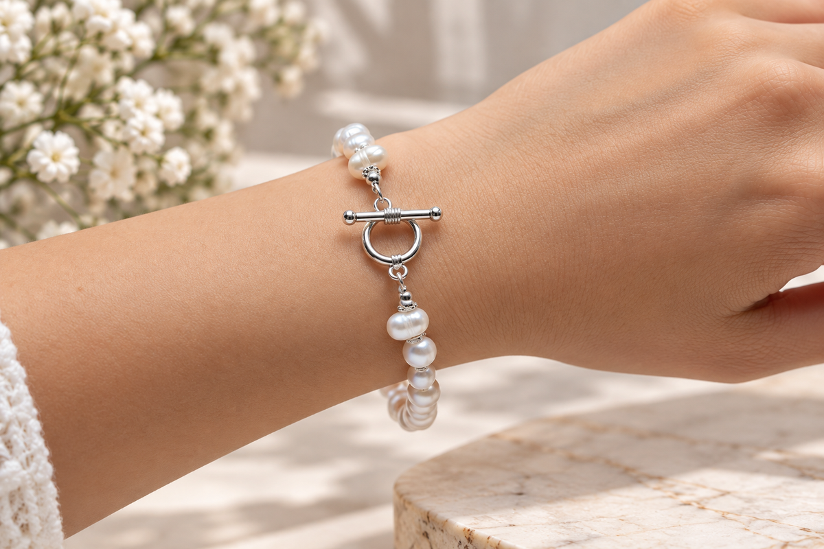 A close-up of a person's wrist wearing a delicate bracelet made of white pearl-like beads and a silver toggle clasp, with soft-focus flowers and a marble surface in the background.