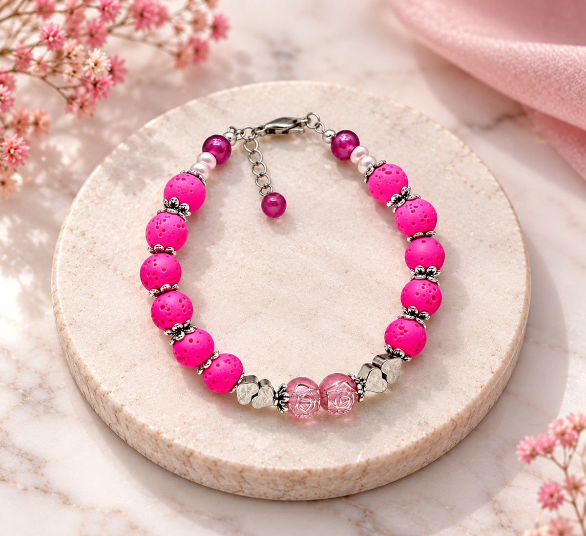 Handmade hot pink lava bead bracelet with silver floral spacers, pink glass beads and heart accents displayed on a round cream stone stand.