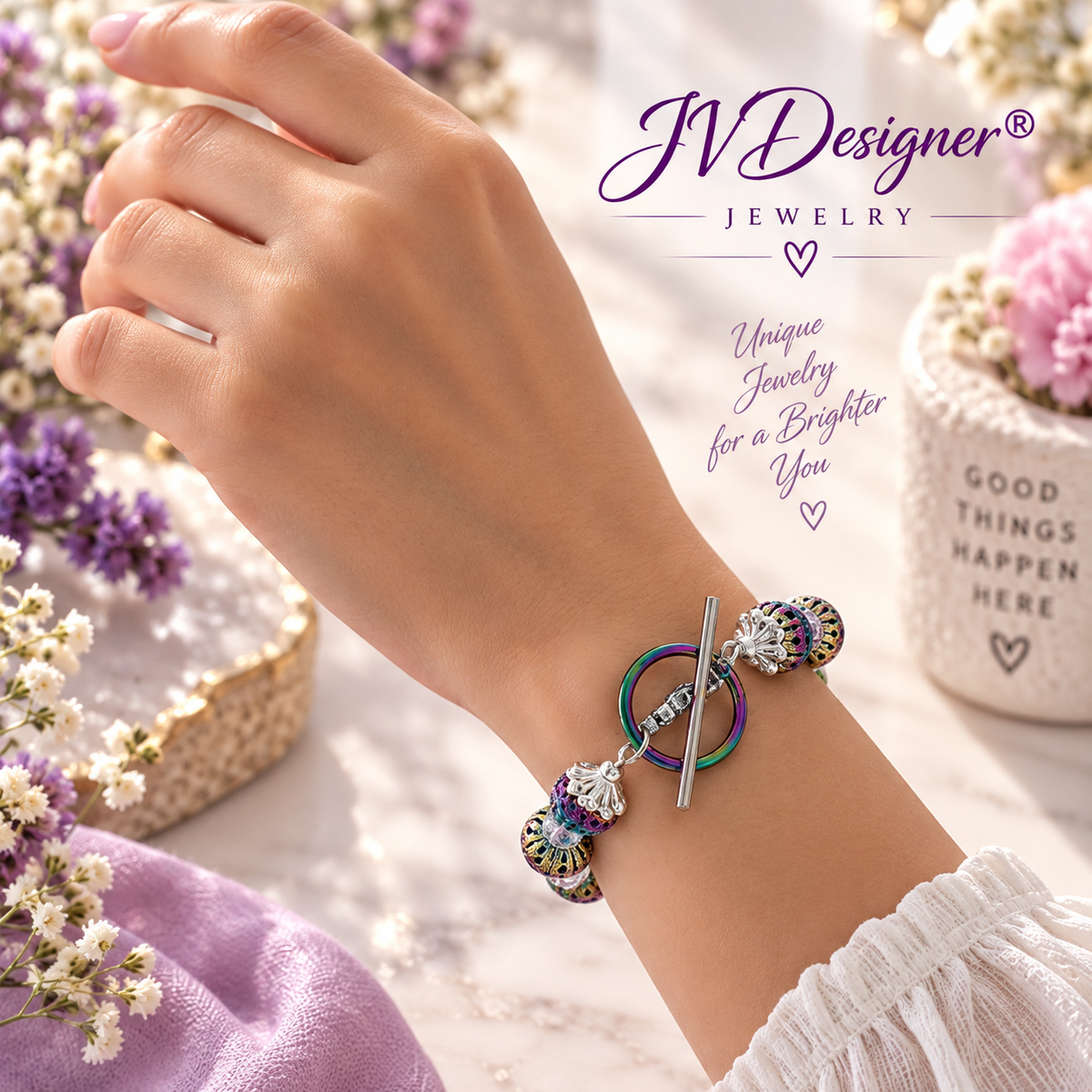 A close-up lifestyle shot of a person's wrist wearing a silver-toned charm bracelet with colorful, iridescent, and floral details. The background is a soft, light marble surface decorated with purple flowers and a small potted plant, with the brand logo 'JVDesigner Jewelry' and promotional text overlaid in the upper right.