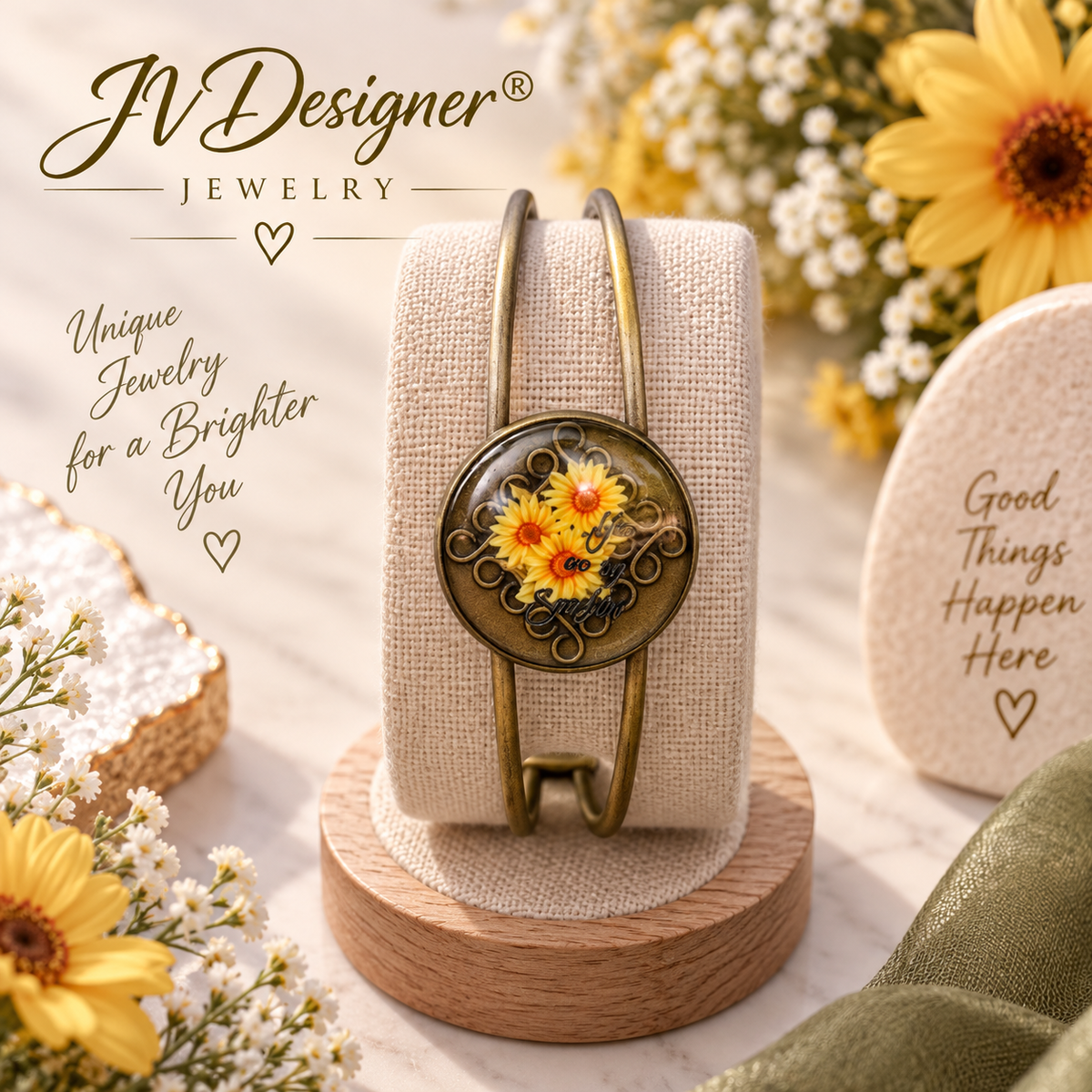 A gold-toned wrap bracelet with a round floral cabochon featuring yellow and orange flowers, displayed on a beige fabric jewelry stand over a wooden base. The scene is styled with sunflowers, white blossoms, and decorative text reading 'JVDesigner Jewelry' and 'Unique Jewelry for a Brighter You'.