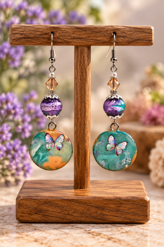 A pair of dangling earrings displayed on a wooden stand. Each earring has a silver fishhook, a faceted clear bead, a purple marbled bead, and a round turquoise and teal disc charm featuring a butterfly design.